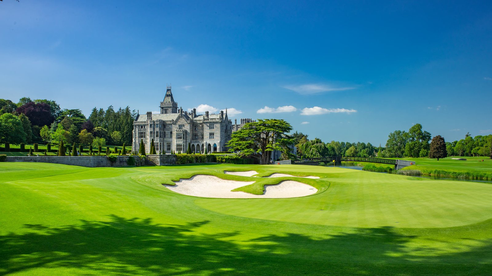 A golf green and white-sand bunkers in front of a large stone manor house and mature trees under a blue sky, Ireland