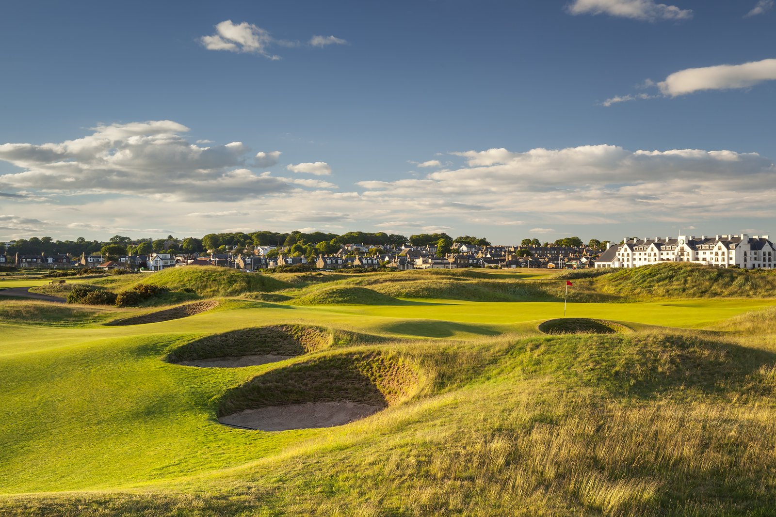 Deep pot bunkers on a sunlit links course, with a Scottish town and a large white hotel on the horizon