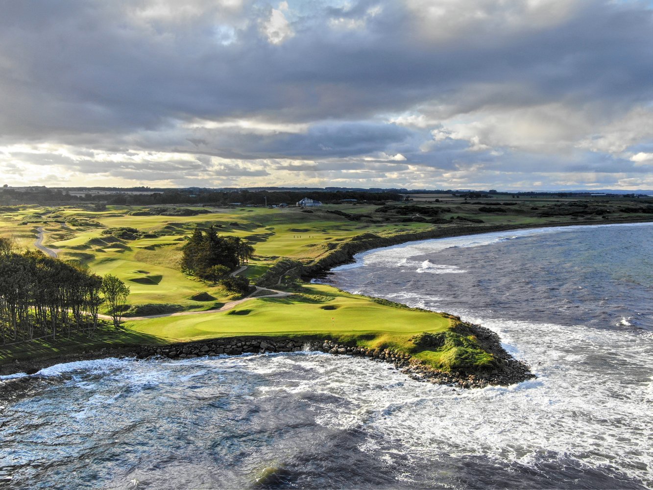 Links holes running along a rocky shoreline with breaking waves and a stand of pines, under a cloudy Scottish sky